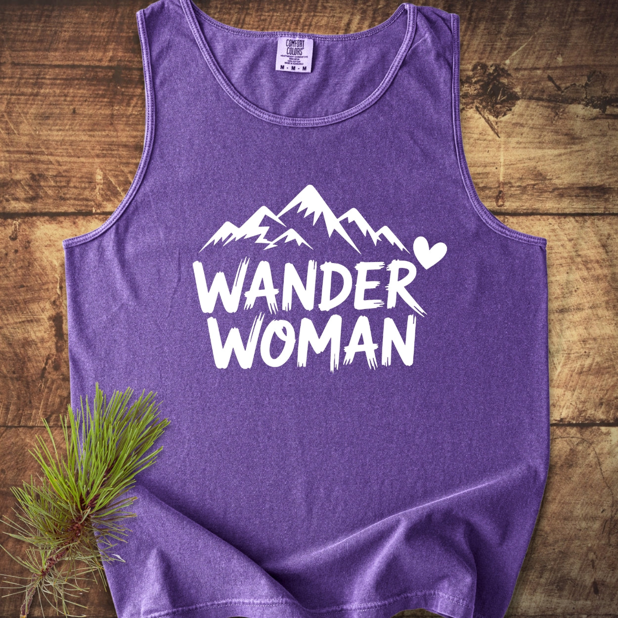 Wander Woman Comfort Colors 9360 Tank Top features a purple tank with a white mountain peaks graphic and 'WANDER WOMAN' text, displayed on a wooden surface with a pine bough accent.