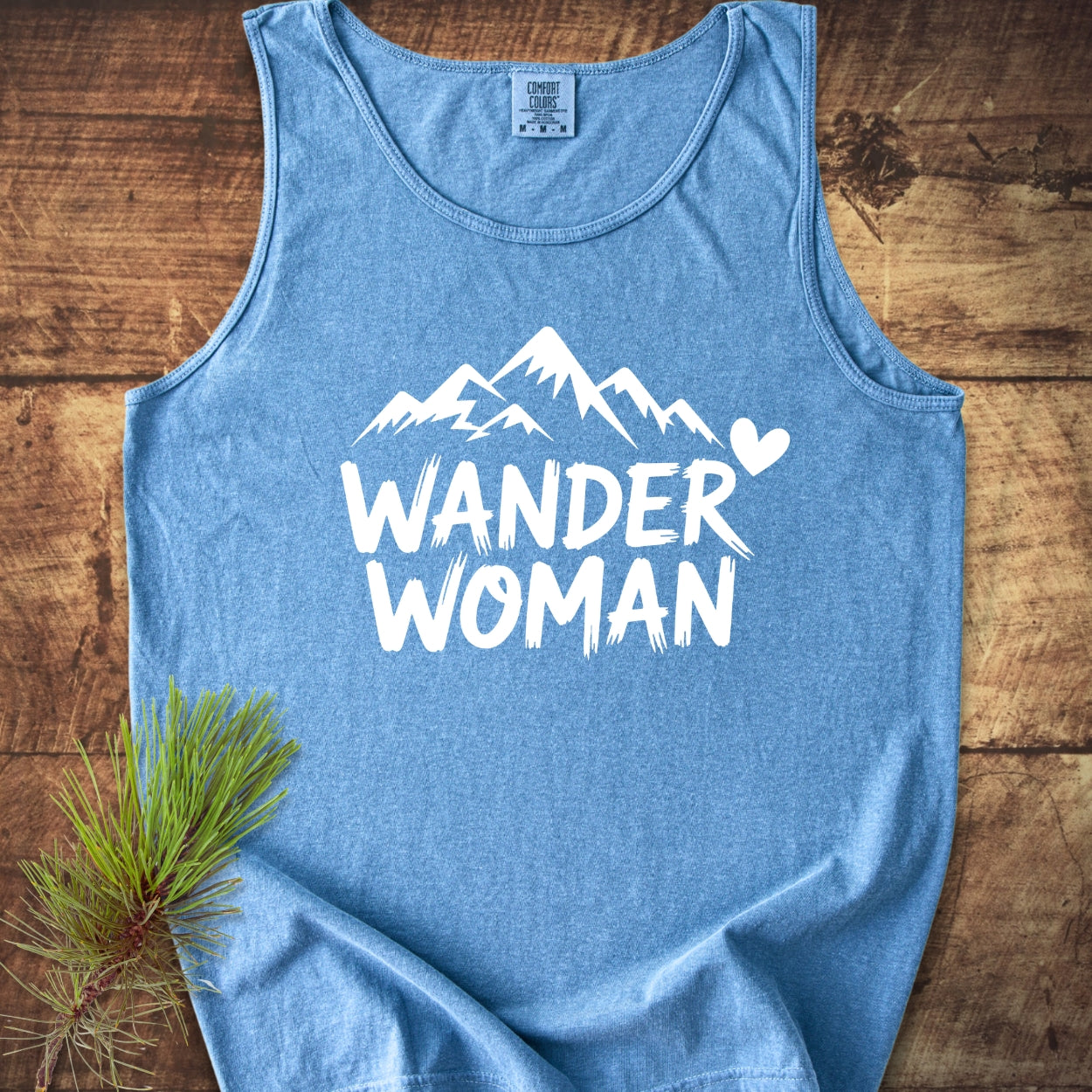 Wander Woman Comfort Colors 9360 Tank Top in heather-blue, featuring a bold white mountain graphic and WANDER WOMAN text, laid on rustic wood with pine sprig, highlighting style and sustainability.