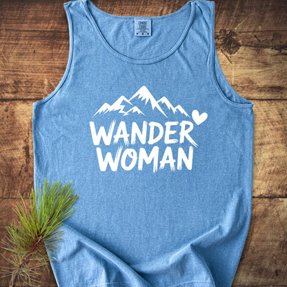 Wander Woman Comfort Colors 9360 Tank Top in heather-blue, featuring a bold white mountain graphic and WANDER WOMAN text, laid on rustic wood with pine sprig, highlighting style and sustainability.