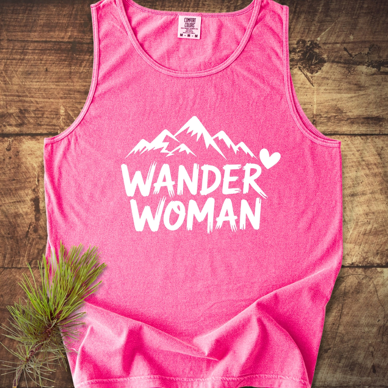 Wander Woman Comfort Colors 9360 Tank Top on rustic wood, featuring a white mountain silhouette and heart design. Beside it, a pine branch highlights its nature-inspired theme, emphasizing sustainability and comfort.