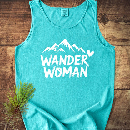 Wander Woman Comfort Colors 9360 Tank Top features a white mountain graphic with WANDER WOMAN text on a turquoise tank, laid on wood with a pine twig for an outdoorsy feel.