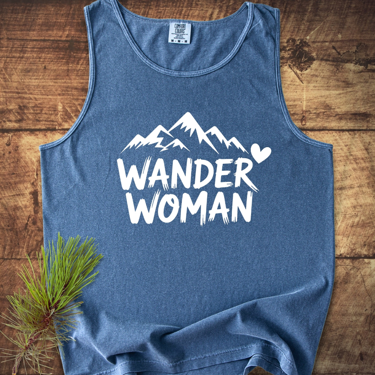 Wander Woman Comfort Colors 9360 Tank Top, a blue sleeveless garment, features a mountain range and heart graphic with WANDER WOMAN text, laid on wood beside a pine branch.