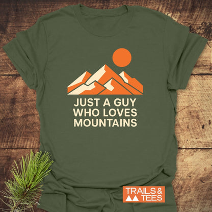 Mountain Love T-Shirt features a stylized mountain and sun print with “JUST A GUY WHO LOVES MOUNTAINS” text, laid flat on wood. Includes “TRAILS & TEES” logo.