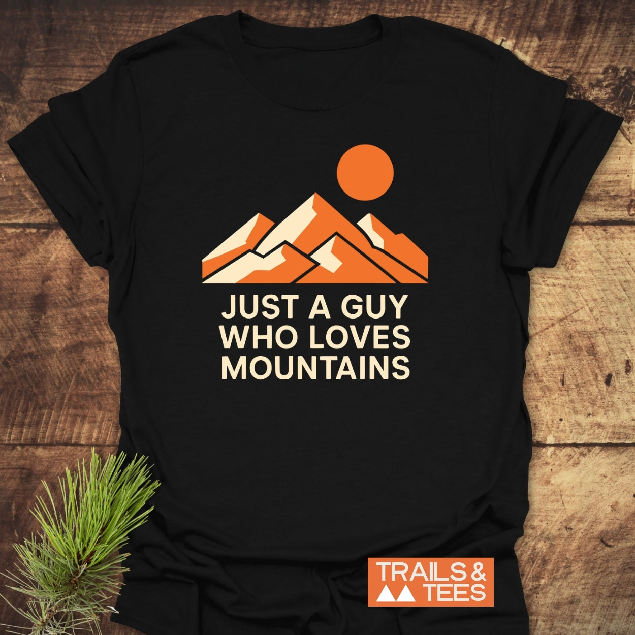 Mountain Love T-Shirt featuring a graphic of three mountain peaks with an orange sun, highlighted by the phrase JUST A GUY WHO LOVES MOUNTAINS. Includes TRAILS & TEES badge.