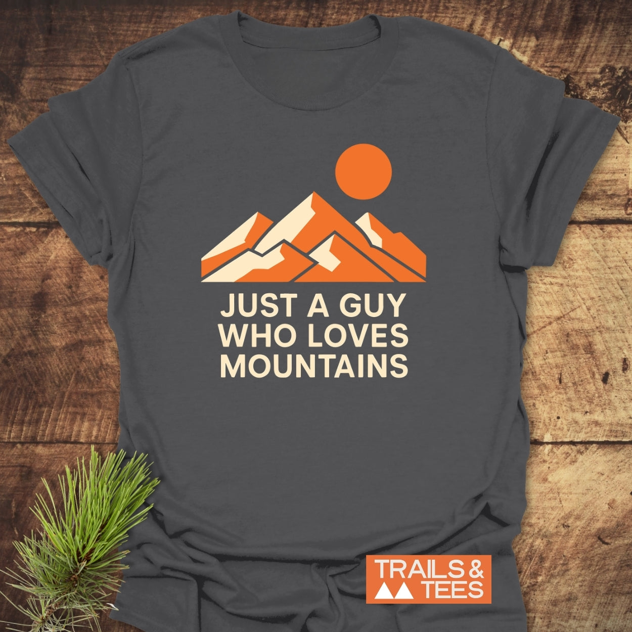 Mountain Love T-Shirt on wooden surface, featuring orange-and-cream mountain graphic with sun and JUST A GUY WHO LOVES MOUNTAINS text. Includes small TRAILS & TEES logo in corner.