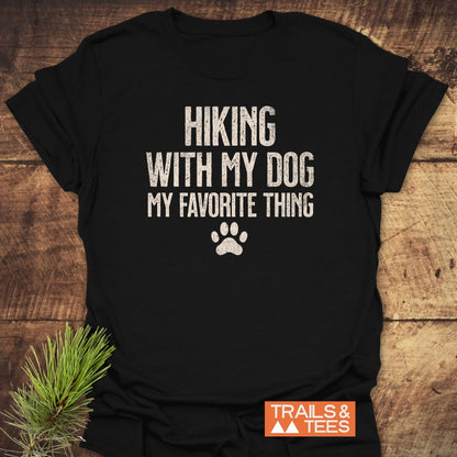 Hiking With My Dog T-Shirt features a distressed print with the phrase HIKING WITH MY DOG MY FAVORITE THING and a paw-print graphic, laid flat with TRAILS & TEES logo visible.