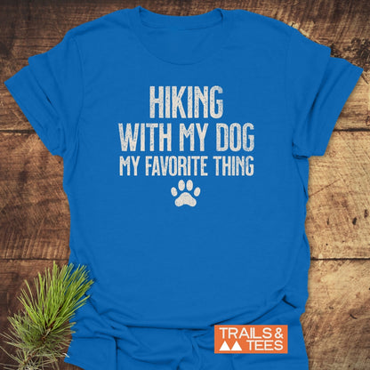 Hiking With My Dog T-Shirt in royal blue with distressed white text and paw print, laid on wood. Features Trails & Tees logo, rustic pine sprig detail.