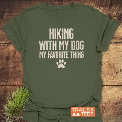 Hiking With My Dog T-Shirt on a wooden surface, featuring HIKING WITH MY DOG MY FAVORITE THING text and paw-print. Includes TRAILS & TEES logo and pine sprigs beside the shirt.
