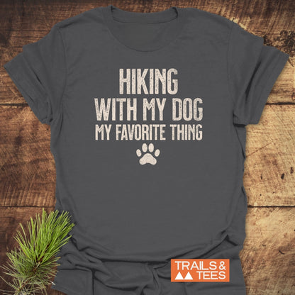 Charcoal-gray Hiking With My Dog T-Shirt featuring distressed white lettering and paw-print, displayed on wood. Includes orange TRAILS & TEES logo and pine branch accent, highlighting outdoor adventure spirit.