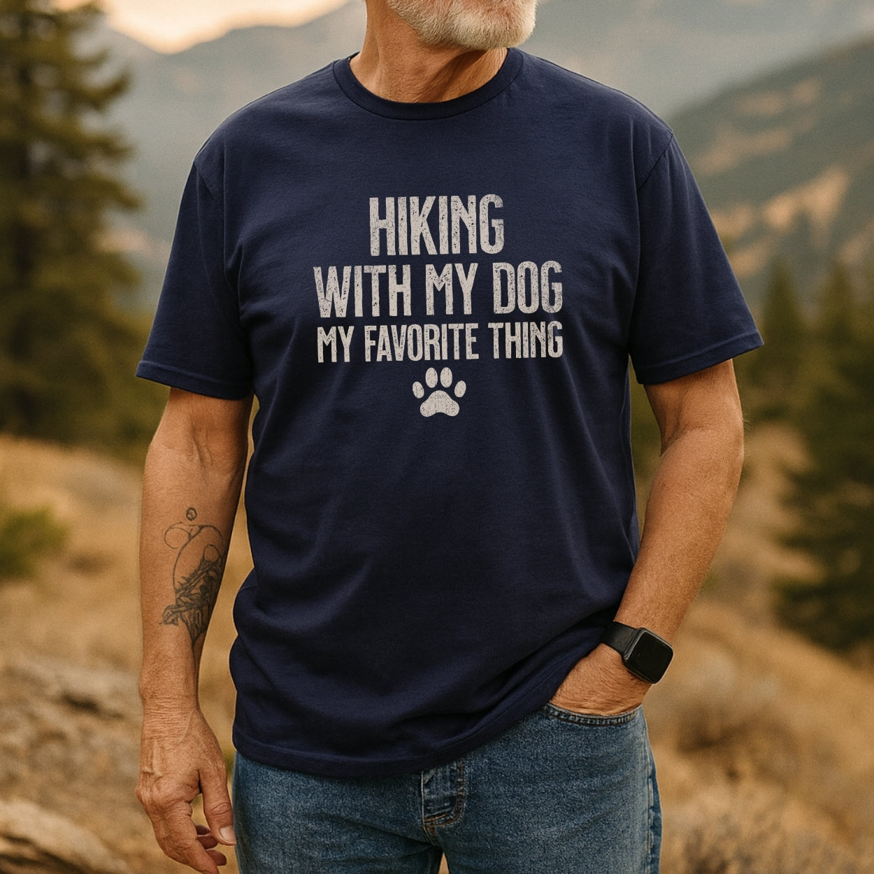 Person outdoors wearing the Hiking With My Dog T-Shirt, displaying white text and a paw-print on navy-blue. Features include a white beard, tattoo, smartwatch, and jeans, set against a nature backdrop.