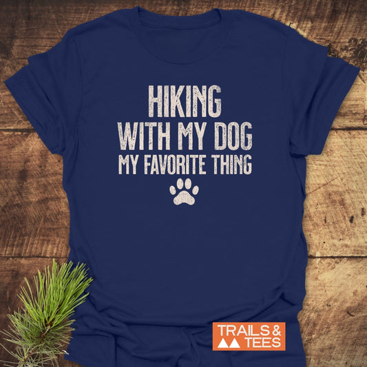 Navy-blue Hiking With My Dog T-shirt with distressed white lettering and paw-print graphic, showcased on a wooden surface with pine sprig, featuring TRAILS & TEES logo in the corner.