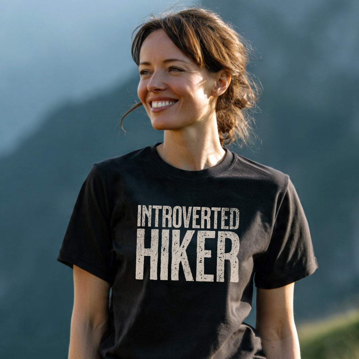 Woman outdoors wearing Introverted Hiker T-Shirt, smiling amidst a natural setting. The dark tee showcases durable DTF print, emphasizing comfort and outdoor spirit with timeless design for adventure lovers.