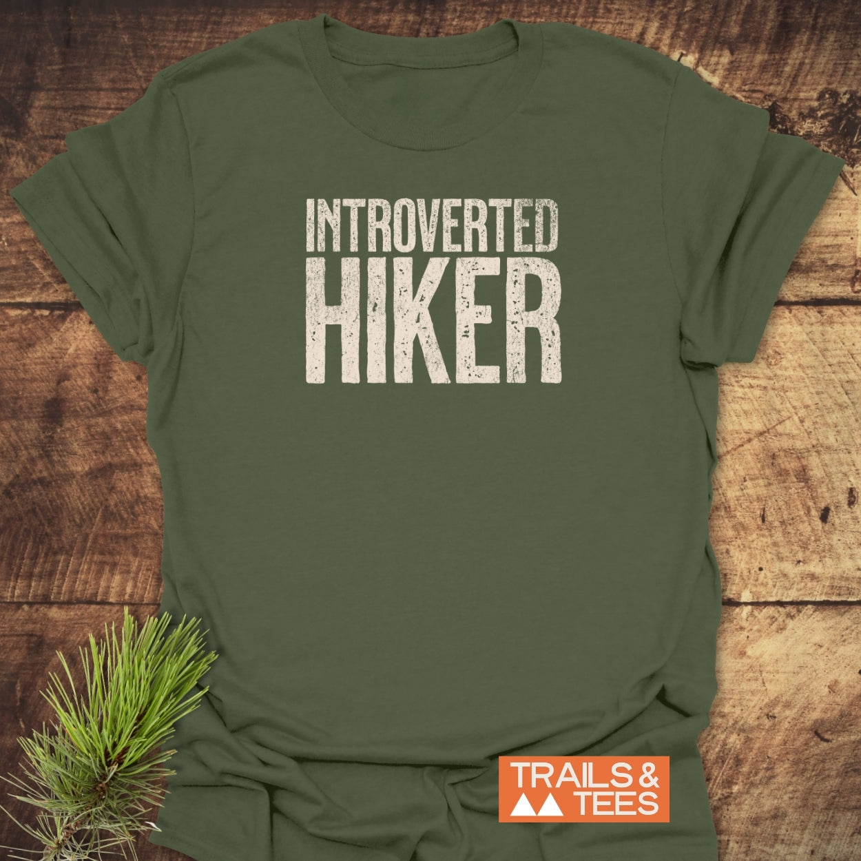 Forest-green Introverted Hiker T-Shirt with a distressed white slogan, featuring a small orange TRAILS & TEES label. Laid on wood with a pine sprig, showcasing outdoor-inspired, comfort-focused design.