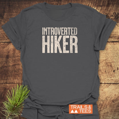 Introverted Hiker T-Shirt on rustic wood, featuring distressed white print. Crafted from soft, lightweight cotton, it embodies outdoor adventure spirit. Branded with Trails & Tees logo and mountain icons.