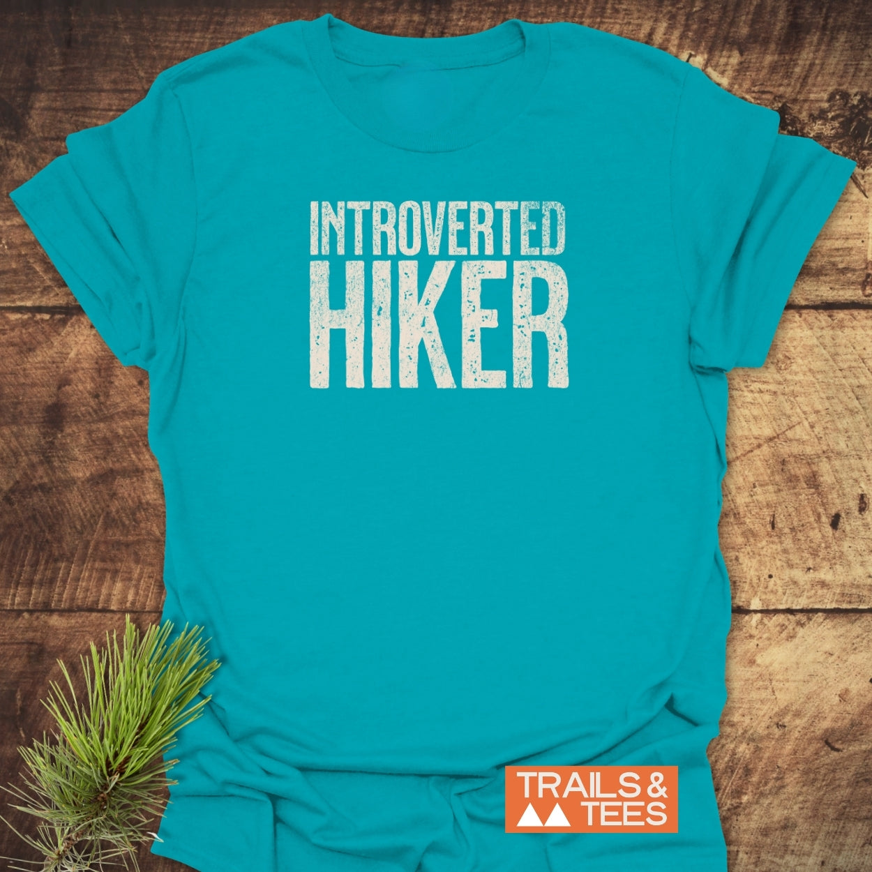 The Introverted Hiker T-Shirt features bold white lettering on soft, lightweight cotton, ideal for outdoor enthusiasts. A rustic pine sprig complements the Trails & Tees logo on a wooden backdrop.