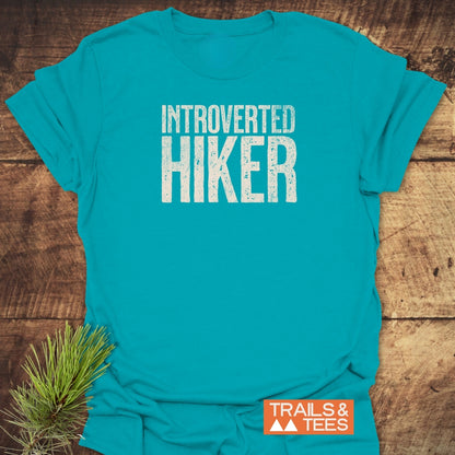The Introverted Hiker T-Shirt features bold white lettering on soft, lightweight cotton, ideal for outdoor enthusiasts. A rustic pine sprig complements the Trails & Tees logo on a wooden backdrop.