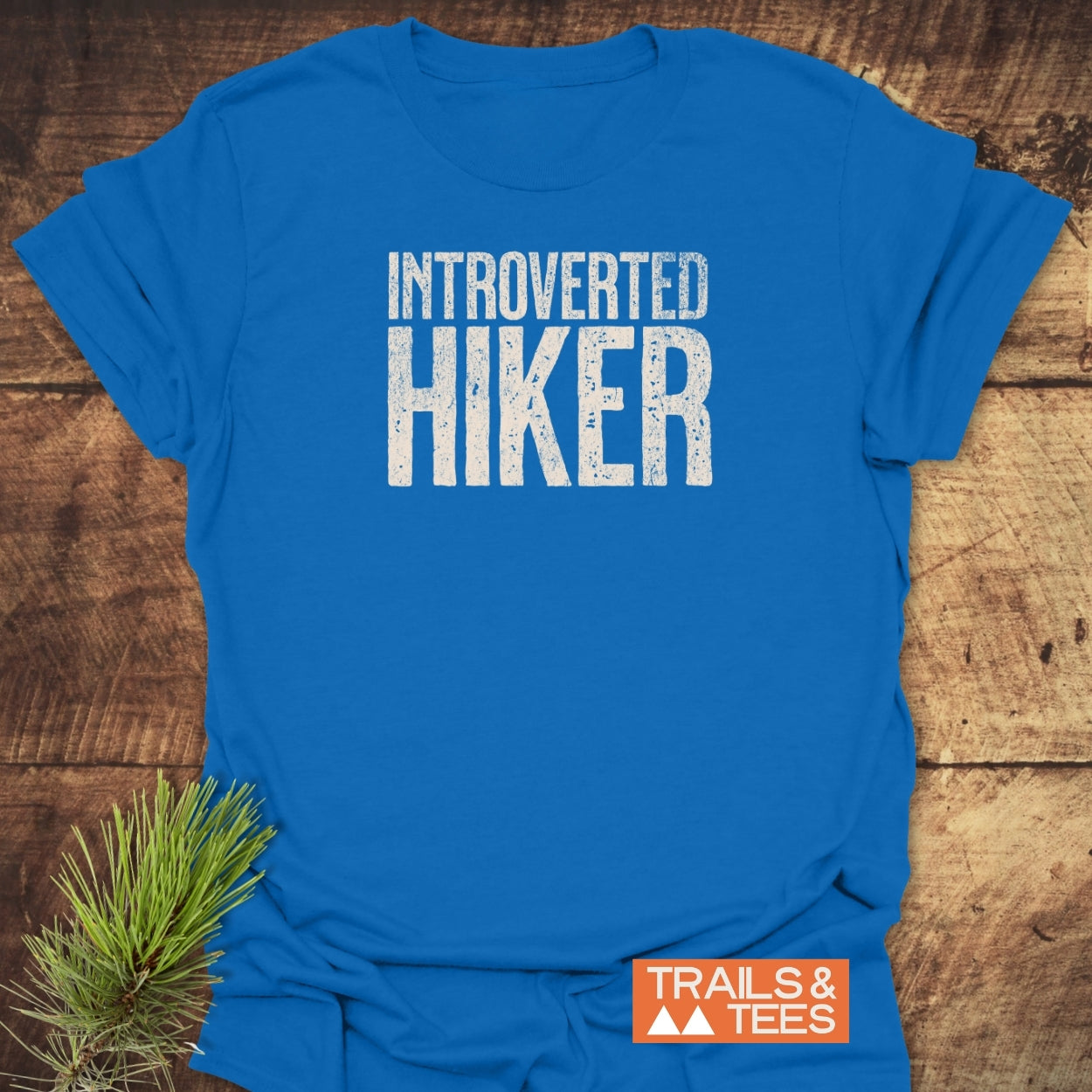 Introverted Hiker T-Shirt displayed on wood-grain surface, featuring weathered white font. Includes TRAILS & TEES logo and pine sprig. Crafted from soft ring-spun cotton for comfort and durability.