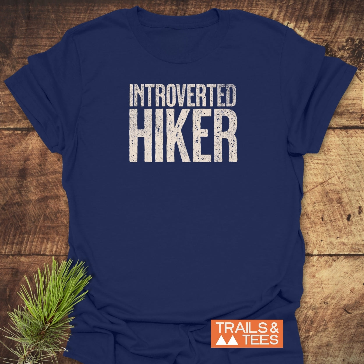 Introverted Hiker T-Shirt on wooden surface, featuring distressed white lettering. Includes “TRAILS & TEES” label with mountain graphic. A pine sprig lies beside, enhancing the shirt's outdoor theme.