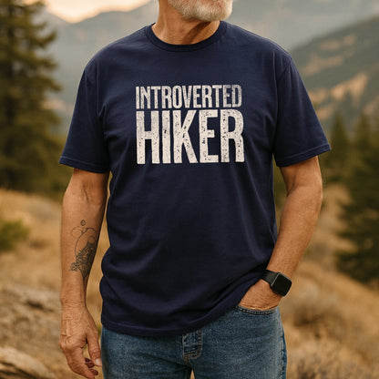 Bearded man wearing an Introverted Hiker T-Shirt stands in a mountain landscape. The navy tee features weathered white lettering, paired with blue jeans and a smartwatch, highlighting outdoor adventure appeal.