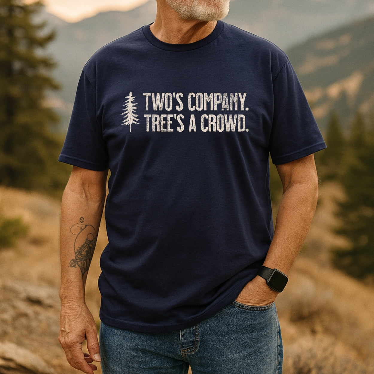 Man in rugged landscape wearing the Two's Company Funny T-Shirt with pine tree graphic and slogan. Paired with jeans, smartwatch, and tattoo, embodying an outdoorsy, adventure-ready vibe.