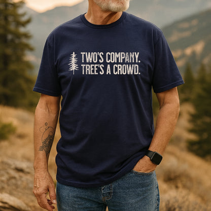 Man in rugged landscape wearing the Two's Company Funny T-Shirt with pine tree graphic and slogan. Paired with jeans, smartwatch, and tattoo, embodying an outdoorsy, adventure-ready vibe.
