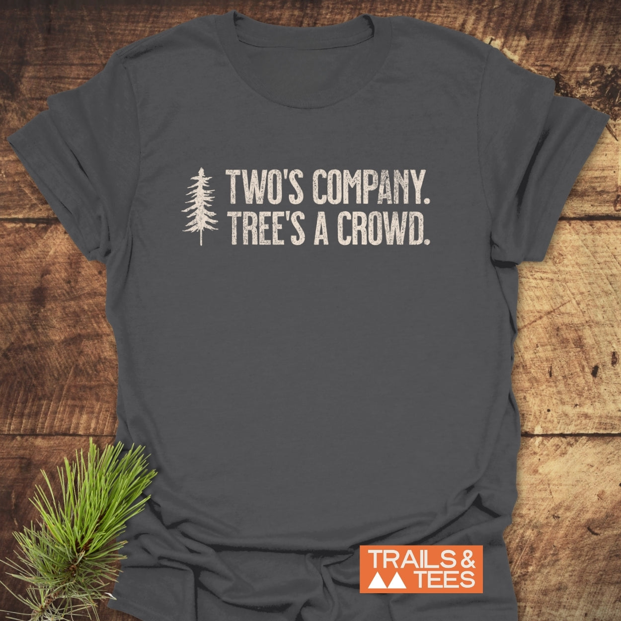 Two's Company Funny T-Shirt featuring a stylized evergreen tree and slogan, laid flat on wood. Crafted from soft ring-spun cotton, it embodies outdoor adventure spirit with durable, distressed white typography.