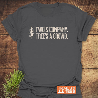 Two's Company Funny T-Shirt featuring a stylized evergreen tree and slogan, laid flat on wood. Crafted from soft ring-spun cotton, it embodies outdoor adventure spirit with durable, distressed white typography.