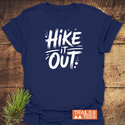 Navy-blue Hike It Out T-Shirt with bold white slogan, displayed on wood. Features TRAILS & TEES orange logo and pine sprig. Crafted for comfort and outdoor enthusiasts, emphasizing durability and sustainability.