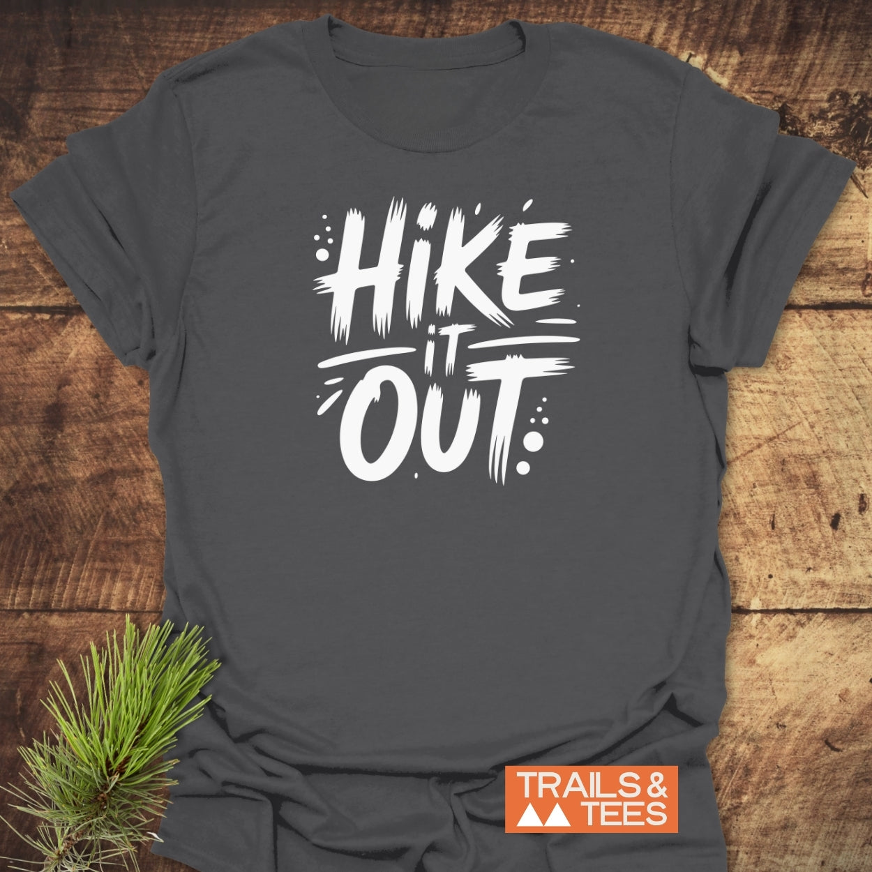 Dark-gray Hike It Out T-shirt with bold white lettering, laid flat with an orange TRAILS & TEES logo, accented by a pine sprig, emphasizing outdoor adventure and comfort.