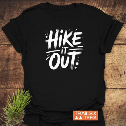 Black Hike It Out T-shirt with bold white slogan, placed on wood. Features an orange TRAILS & TEES logo and pine sprig, highlighting its rustic, outdoor-inspired design.