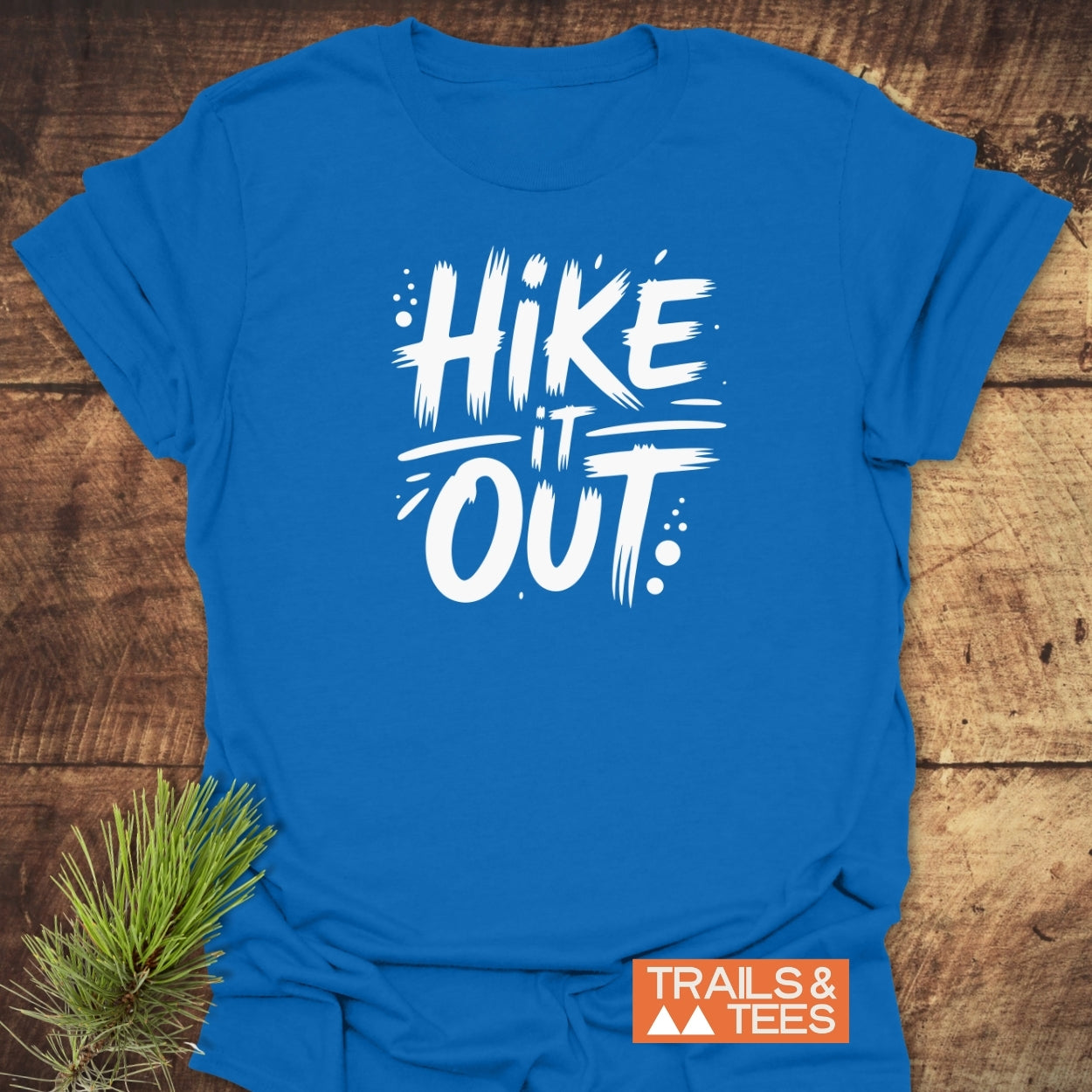 Hike It Out T-Shirt featuring a white brush-stroke slogan on a blue tee, laid flat with a pine sprig. Ideal for outdoor enthusiasts, symbolizing comfort and adventure.