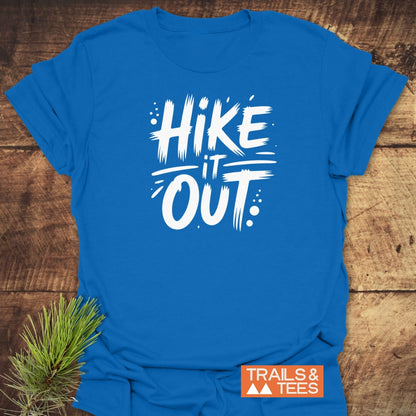 Hike It Out T-Shirt featuring a white brush-stroke slogan on a blue tee, laid flat with a pine sprig. Ideal for outdoor enthusiasts, symbolizing comfort and adventure.