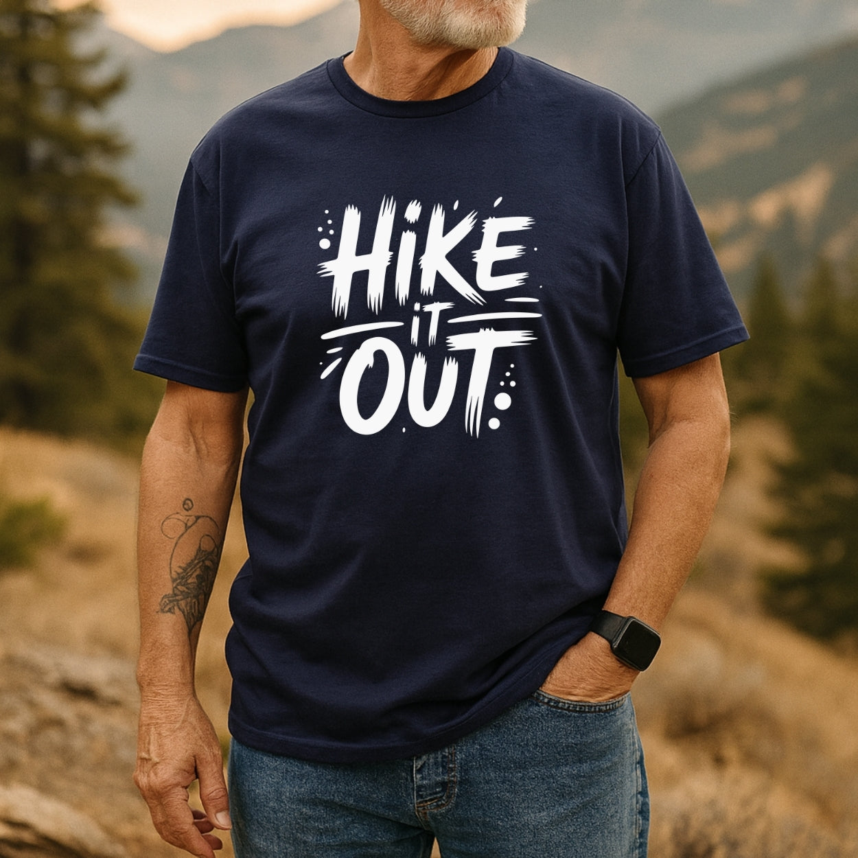 Person outdoors wearing the Hike It Out T-Shirt with bold white lettering, paired with jeans and a wristwatch, showcasing a tattooed forearm against a scenic backdrop of hills and trees.
