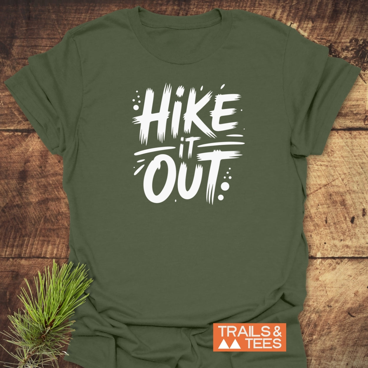 Olive-green Hike It Out T-shirt with bold white slogan, laid flat on wood, featuring TRAILS & TEES orange logo and pine sprig, showcasing trail-ready comfort and adventure spirit.
