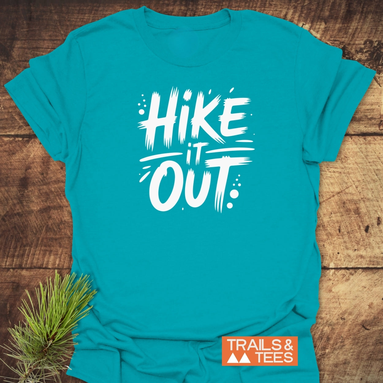 Hike It Out T-Shirt featuring a bold white brush-style slogan on teal, laid flat with a pine sprig alongside, symbolizing an outdoor theme. Includes Trails & Tees logo in the corner.