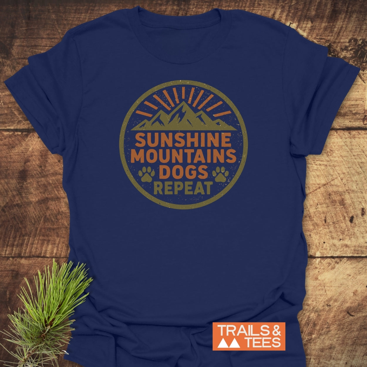 Sunshine Mountains Dogs Badge T-Shirt featuring a circular graphic with mountains, sunbeams, and paw prints. Navy-blue, soft ring-spun cotton, laid flat on wood with a TRAILS & TEES logo.