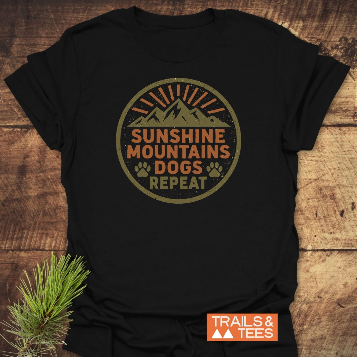 Sunshine Mountains Dogs Badge T-Shirt features a circular graphic with mountains, sun-rays, and paw prints, laid on rustic wood, highlighting a small TRAILS & TEES logo and pine sprig accent.