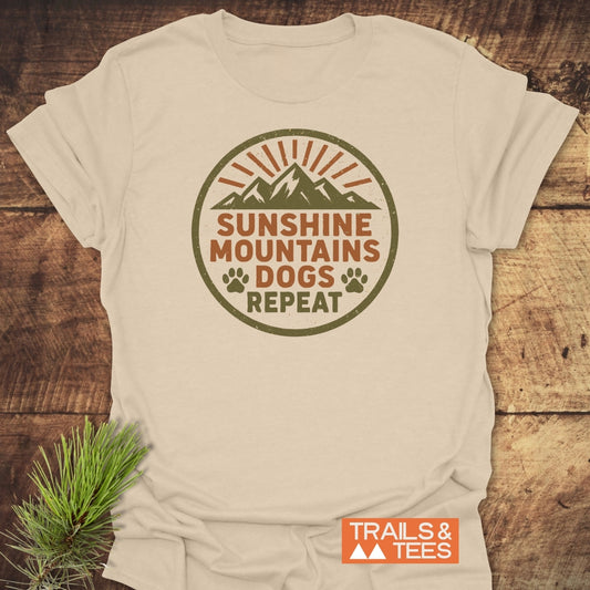 Sunshine Mountains Dogs Badge T-Shirt displayed on wood, featuring a mountain logo with sun rays, paw prints, and slogan. Includes TRAILS & TEES mark. Pine sprig adds an outdoor touch.