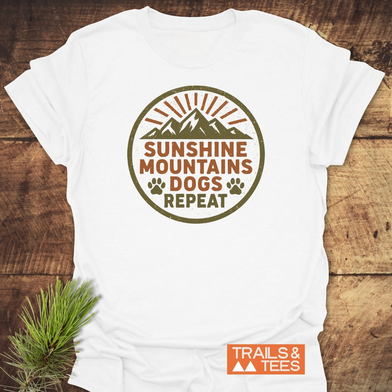 Sunshine Mountains Dogs Badge T-Shirt on wood surface; features a graphic with mountain peaks, sun rays, paw prints, and slogan. Includes TRAILS & TEES logo, highlighting outdoor adventure theme.
