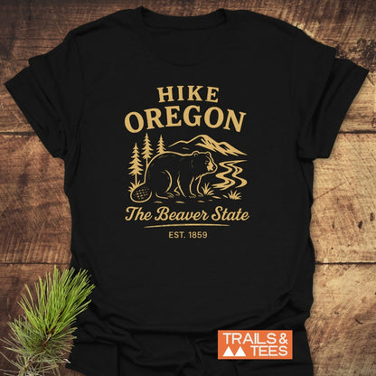 Hike Oregon T-Shirt featuring a gold beaver graphic with mountains and trees, laying on wood. Includes The Beaver State EST. 1859 text, conveying an outdoor adventure theme.