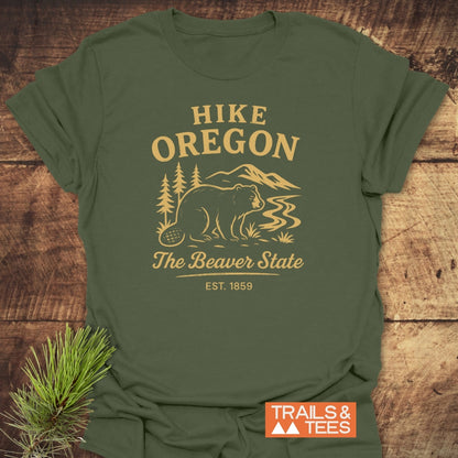 Hike Oregon T-Shirt featuring a golden-tan beaver scene with The Beaver State and EST. 1859 text, laid on wood with a pine branch, highlighting outdoor adventure spirit.
