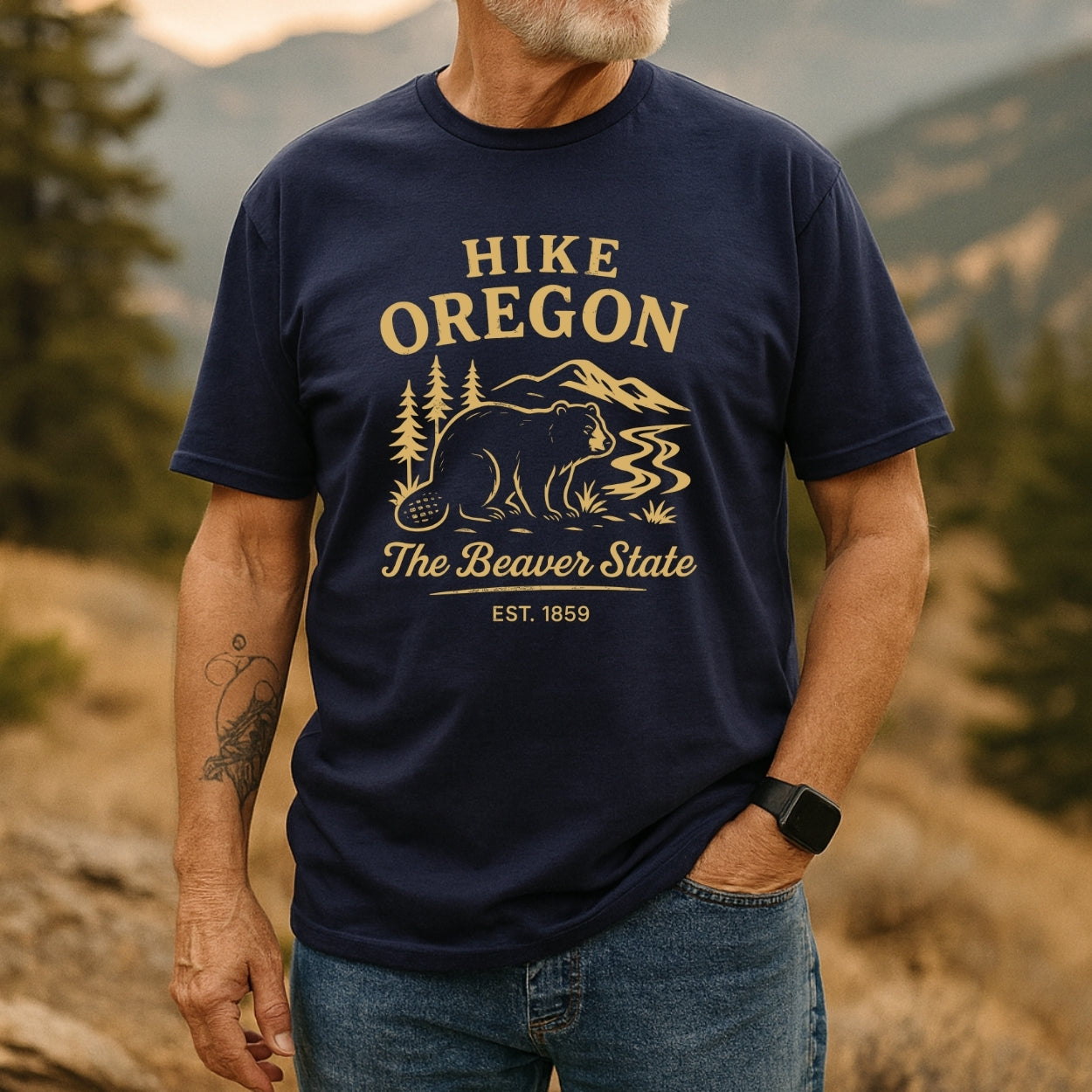 Man wearing the Hike Oregon T-Shirt with a beaver, pine trees, and mountain graphic, styled with jeans and smartwatch, embodying outdoor adventure spirit in a mountainous, forest-edge setting.