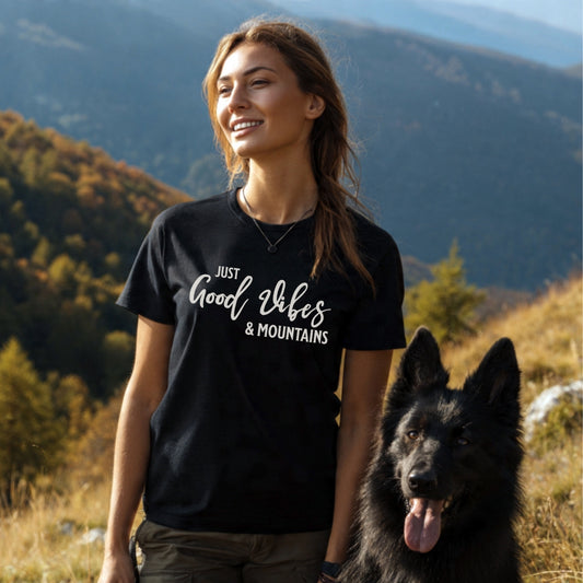 Young woman in a Just Good Vibes & Mountains T-Shirt stands with a large black dog in a hilly, forested autumn landscape, highlighting the tee's outdoor adventure spirit and comfort.