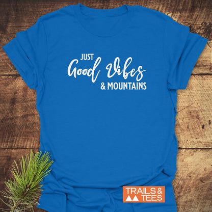 Just Good Vibes & Mountains T-Shirt on rustic wood, featuring white script and block letters. Includes the “TRAILS & TEES” logo with mountain icons, complemented by a sprig of pine needles.