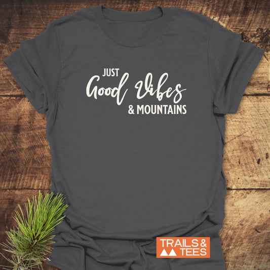 Just Good Vibes & Mountains T-Shirt features white lettering on dark-gray fabric, showcasing an orange TRAILS & TEES logo and a pine sprig, embodying outdoor adventure and comfort.
