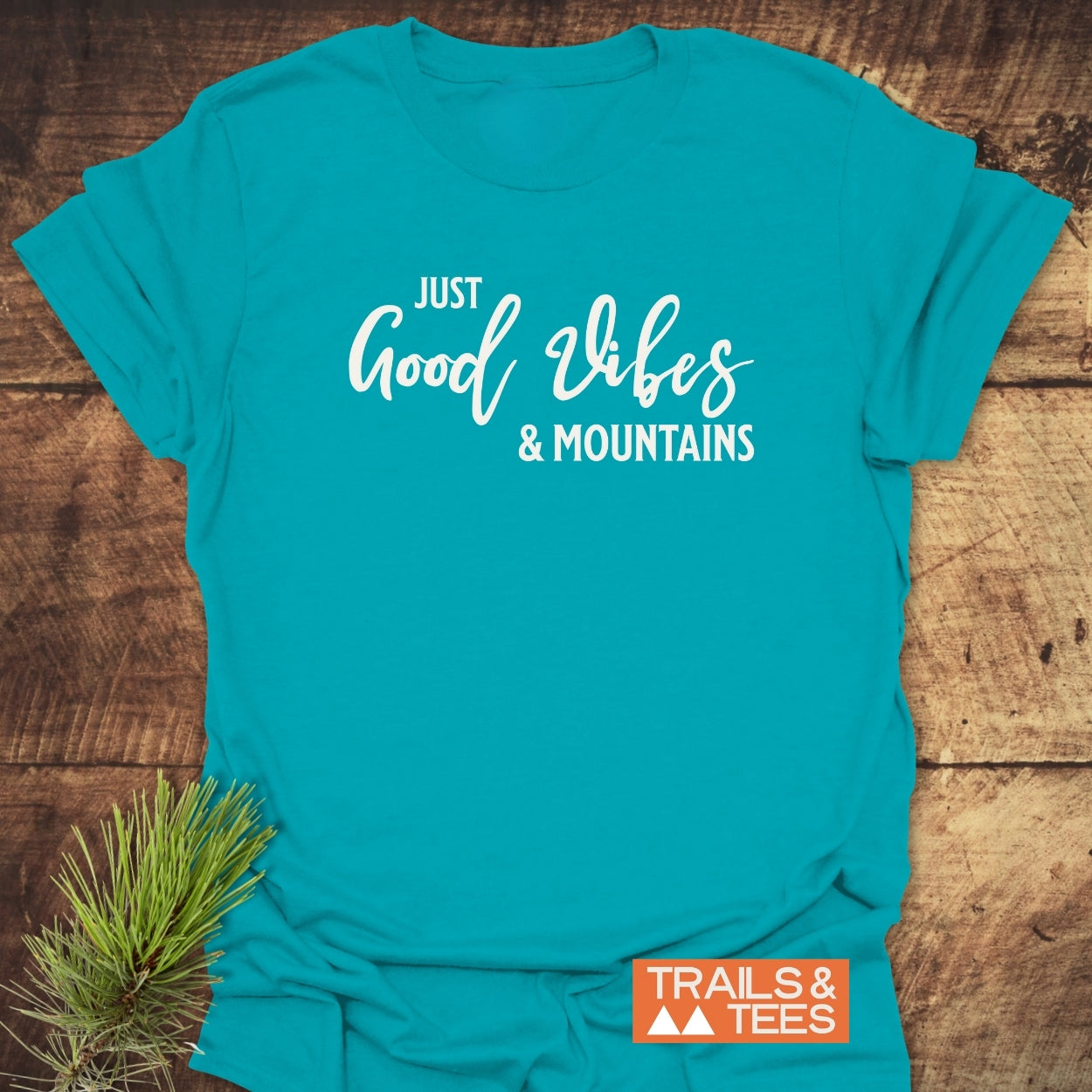 Just Good Vibes & Mountains T-Shirt laid flat, showcasing a white slogan on a teal crew-neck. Includes TRAILS & TEES logo and pine needles, emphasizing outdoor adventure spirit with durable, comfortable design.