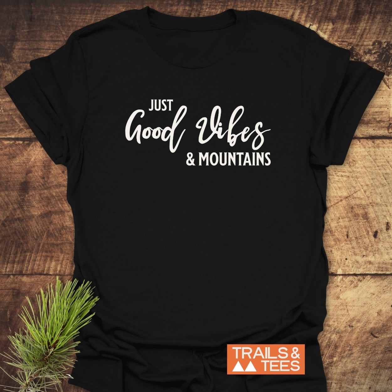 Black Just Good Vibes & Mountains T-shirt on wood, featuring white lettering. Includes Trails & Tees logo and pine sprig. Crafted from soft ring-spun cotton for outdoor enthusiasts.