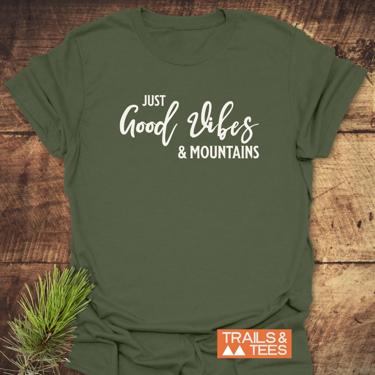 Just Good Vibes & Mountains T-Shirt, olive-green, with white text, laid on rustic wood. Includes pine sprig accent, orange TRAILS & TEES logo, embodying outdoor adventure and comfort.