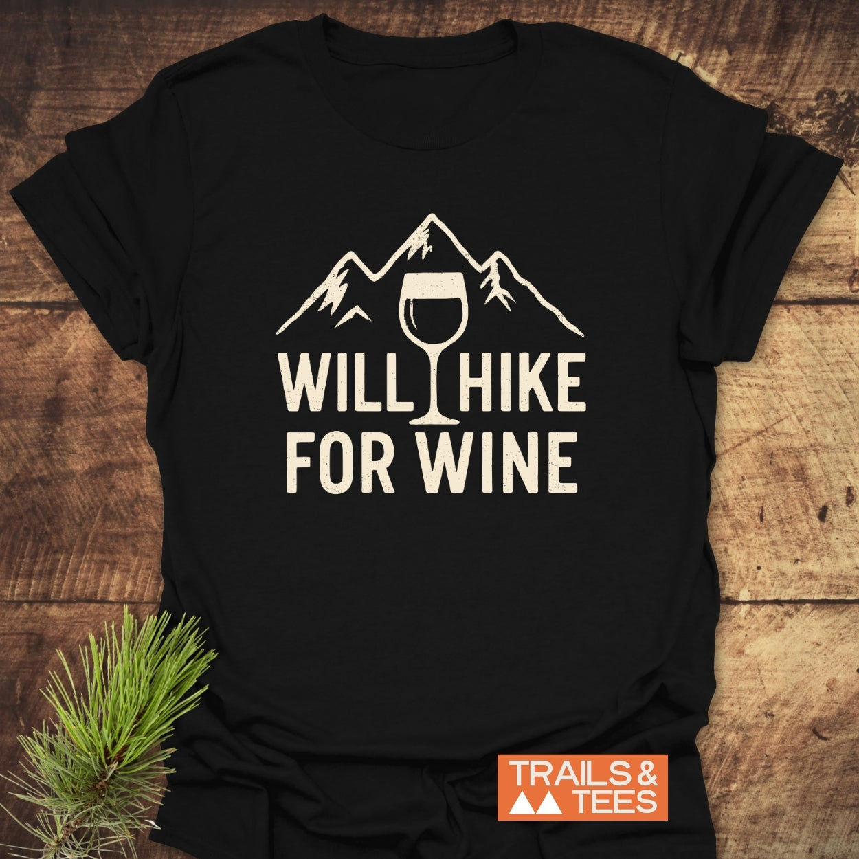 Will Hike For Wine T-Shirt featuring a mountain and wine glass design, laid on wood. Crafted from soft cotton, it offers trail-tested comfort with durable prints and a small Trails & Tees logo.