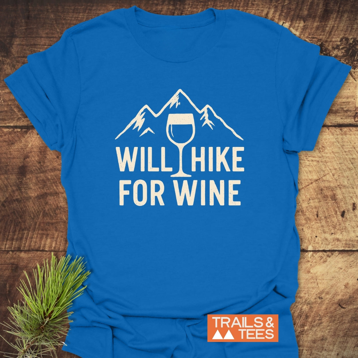 Will Hike For Wine T-Shirt with mountain and wine glass design, laid on wood surface. Features soft ring-spun cotton, durable print, and Trails & Tees logo, embodying outdoor adventure spirit.